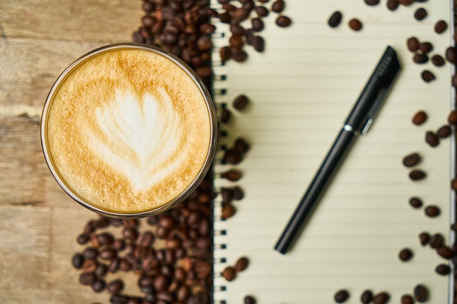 A top view of a latte next to a notebook with a pen, with coffee beans strewed artfully around.